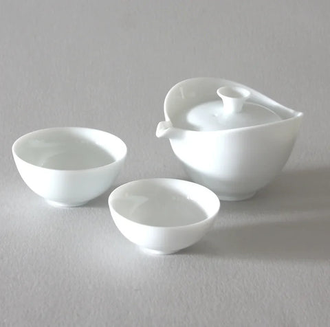 The Ingot White Travel Tea Set by Eastern Elm is a high-quality ceramic tea set that includes a teapot with a spout and lid, accompanied by two small teacups. The entire set is displayed on a light gray surface, showcasing its minimalist and elegant design.