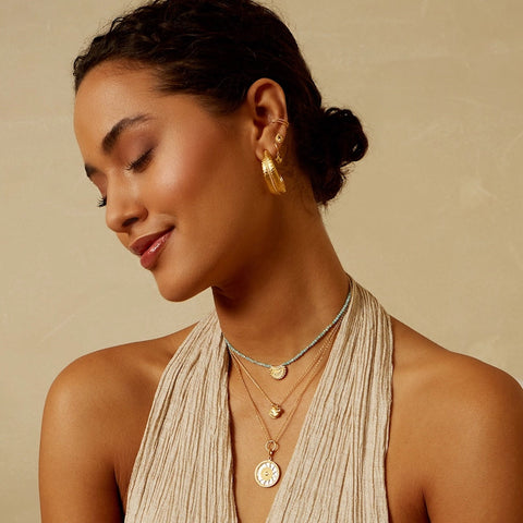 A woman with dark hair in a low bun smiles with her eyes closed, showcasing gold hoop earrings and multiple layered 18KT gold plate necklaces, including the stunning Inward Journey Pearl Mandala Necklace. She is wearing a light beige, sleeveless top and stands against a neutral background.