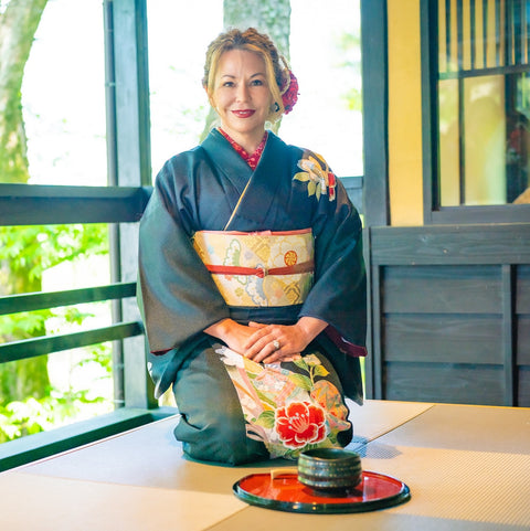 A woman kneeling on a tatami mat, adorned in a traditional black kimono with floral patterns and a colorful obi, smiles serenely in a Japanese-style room with sliding doors and views of a garden. In front of her lies the Tea School 2024-2025 Full Course Bundle by Magic Hour, displayed on a red lacquer tray with stacked bowls, echoing the traditions of the Japanese Tea School.