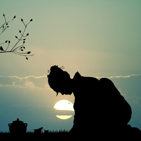 Silhouette of a person in traditional clothing bowing with the setting sun behind them. To the left, a branch with leaves extends into the scene, while a teapot and cup filled with Jasmine Moon White Tea from Magic Hour are positioned on the ground on the right side of the image.