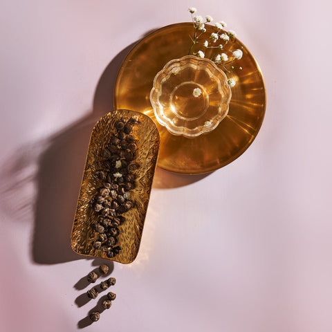 A flat lay image displaying a brass serving tray with dried spices, a glass bowl with a brass base holding a beverage, and a small sprig of Dragon Phoenix Jasmine Pearls by Magic Hour. The items are arranged on a soft pink surface, casting gentle shadows.