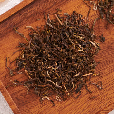 A close-up view of a pile of Jasmine Yin Hao Green Tea by Magic Hour on a wooden surface. The tea leaves are loosely spread out, showcasing their dark, twisted, and curled textures. The wooden background adds a natural, rustic feel to the image.