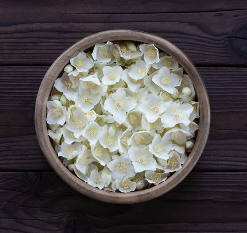 A wooden bowl brimming with delicate magnolia oolong jasmine flowers rests on a dark wooden surface. The white petals create a striking contrast against the rustic background, evoking the sophisticated elegance celebrated by Magic Hour's Triple Moon Tea by Alanis Morissette for her Triple Moon Tour.