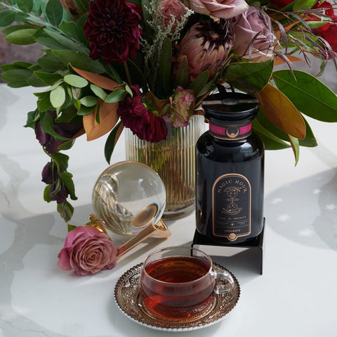 A colorful floral arrangement sits in a vase on a table next to a bottle labeled "Magic Hour" and a teacup filled with Soulmate: Chocolate-Raspberry-Rose Black Tea for Finding & Celebrating Love by Magic Hour. A glass orb and a single pink rose are also on the table, creating an elegant and inviting scene.