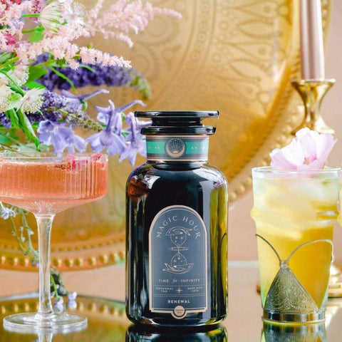 A visually appealing arrangement featuring a dark glass bottle of Magic Hour Renewal: Peach-Goji-Rose Oolong Tea placed on a reflective surface. It is flanked by two ornate glasses containing colorful beverages, with a bouquet of flowers and a decorative gold background enhancing the scene, highlighting the essence of loose leaf tea.