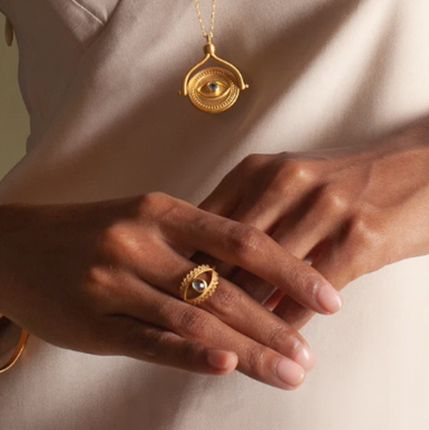 Close-up of a person's hands wearing the Keeper of Positivity Eye Blue Topaz Ring, featuring an eye-shaped design with a blue topaz at its center. The person's chest displays a matching gold necklace with a similar evil eye pendant. The background is softly lit, highlighting the jewelry and the person's light, neutral-colored clothing.