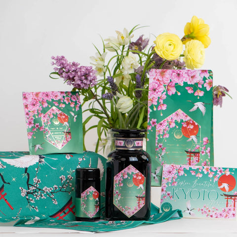 An arrangement of Queen of Kyoto Case Packs by Magic Hour, showcasing floral packaging inspired by cherry blossoms and Kyoto. The collection features jars, pouches, and boxes adorned with pink flowers. Highlighting the Japanese Cherry Blossom Festival, the display includes a bouquet of assorted flowers in a vase as the backdrop.