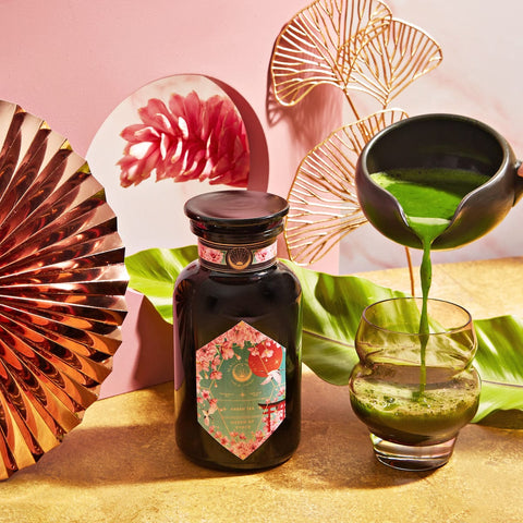 A vibrant setup featuring a jar of Queen of Kyoto Case Packs matcha powder by Magic Hour with an ornate label, surrounded by elegant decorations including a copper fan, tropical leaf, and abstract pink art. A hand holds a black teapot, pouring matcha tea into a clear glass on a gold surface, reminiscent of the Japanese Cherry Blossom Festival.