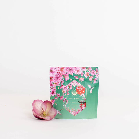 A green package adorned with pink floral designs and decorated with a transparent window revealing its contents. It is labeled with the "Magic Hour" brand and named "Queen of Kyoto Case Packs," featuring organic ingredients. A pink and white orchid flower sits beside the package against a plain white background.