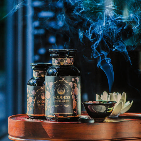 Two dark glass jars labeled "Goddess of Earl: Lady Luck Oriental Beauty Oolong Tea" from Magic Hour are placed on a wooden surface. A small bowl releases smoke, contributing to the serene ambiance. The background is softly blurred, accentuating the aromatic setting of this organic loose leaf tea captured in the image.