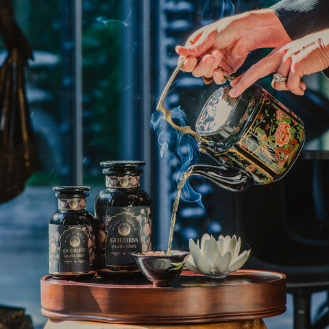 A person pours organic tea from an ornate teapot into a small cup. The table features two black jars labeled "Goddess of Earl: Lady Luck Oriental Beauty Oolong Tea" by Magic Hour. A white lotus-shaped ornament is also on the table. Steam rises from the cup, suggesting the tea is hot, adding to the serene magic hour of tea time.