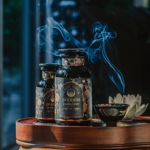 Two dark glass bottles of "Goddess of Earl: Lady Luck Oriental Beauty Oolong Tea," a Magic Hour product, sit on a wooden tray adorned with intricate labels. Wisps of pale smoke rise from a ceramic dish with incense next to a white flower, creating a tranquil and aromatic atmosphere, perfect for savoring this organic tea.