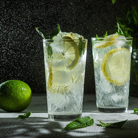 Two glasses of Magic Hour's Lemon Lime Green Tea, a ceremonial soda pop tea with Sugar "Destroyer" and organic ingredients, garnished with slices of lemon and mint leaves, sit on a textured surface with water droplets shimmering in the background. A whole lime and scattered mint leaves are in the foreground.