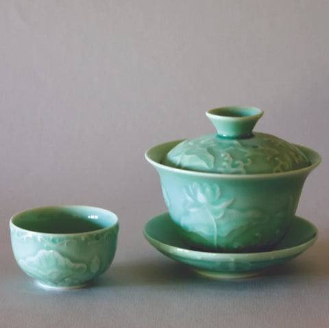 The Lotus Gaiwan Tea Set by Eastern Elm features a jade-green porcelain tea set that includes a covered gaiwan on a saucer and a matching teacup. Delicate floral motifs and intricate designs celebrate Chinese tradition, set against a plain background. The lustrous glaze enhances its elegant appearance.