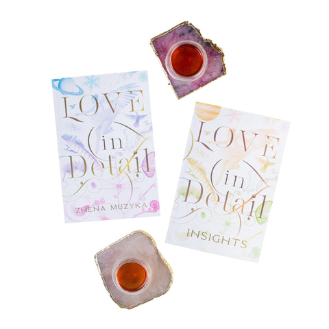 Two copies of "Love in Detail" by Zhena Muzyka are laid out on a white background. One book is paired with a smaller booklet titled "Insights," which is ideal for spiritual guidance. The display also features two crystal cup holders, each containing a small cup filled with reddish liquid.