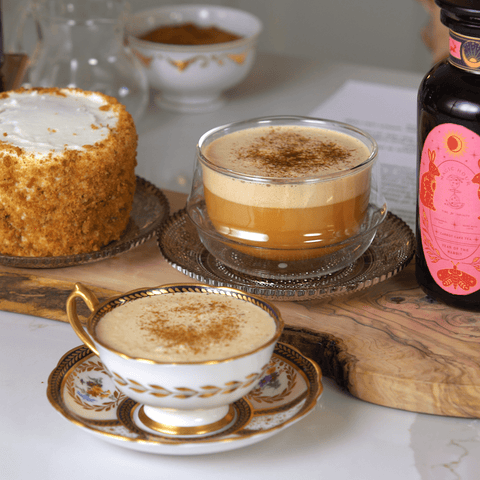 A beautifully arranged coffee setup featuring a vintage teacup filled with frothy Year of the Rabbit Carrot Cake Tea, a glass cup of cappuccino topped with cinnamon, a bottle with a pink label, and a small round cake with white frosting on a decorative tray.