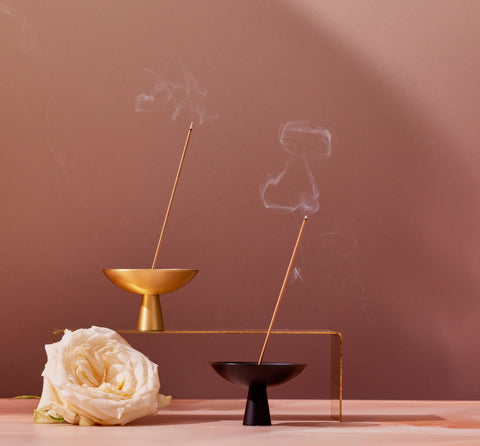 A minimalist scene featuring Magic Hour's high-quality Ceremonial Incense Burners in brass and black, with lit Japanese incense sticks emitting smoke. The brass holder is elevated on a metal stand. A single white rose lies beside them against a neutral tan background.