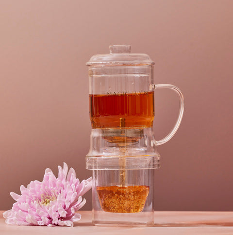 An elegant design, the clear Glass Tea Infuser by Alibaba includes a lid and sits atop the matching glass Magic Hour Glass Tea Infuser, allowing tea to seep from the pot above into a lower container. To the left of this premium quality setup, a pink chrysanthemum flower rests on the surface. The background is a warm, light brown color.