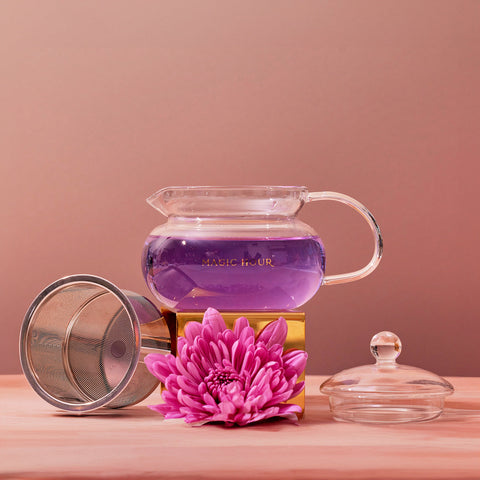 A handcrafted 450ml Magic Hour Glass Teapot filled with purple liquid is set on a wooden surface. Beside it, there is a metal mesh tea infuser, a vibrant pink flower, and a glass lid. The background gradient transitions from peach to mauve, complementing the elegant setup.