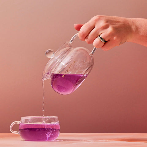 A hand wearing rings pours vibrant purple tea from a clear Magic Hour Glass Teapot into a matching glass teacup against a soft, peach-toned background.