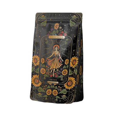 A vibrant black package adorned with detailed illustrations of sunflowers and leafy vines, featuring a central image of a woman in a flowing dress amidst sunflowers. Text reads "Magic Hour" and "The Sun: Energizing Adaptogenic Herbal Tea with Yaupon, Moringa & Turmeric," hinting at the natural blend within.