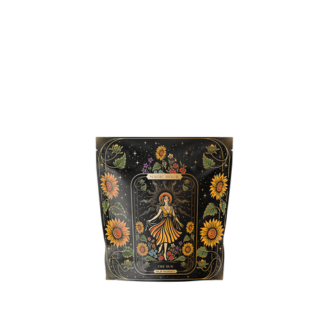 A black packaging bag with floral designs, featuring a central tarot card illustration labeled "The Sun." The design includes a crowned figure surrounded by sunflowers and celestial motifs. Notably, this Magic Hour blend, named "The Sun: Energizing Adaptogenic Herbal Tea with Yaupon, Moringa & Turmeric," contains rejuvenating ingredients like Moringa and Turmeric Root.