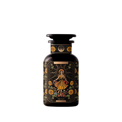 A dark amber bottle from Magic Hour, adorned with ornate label designs, features an illustration of a woman in a yellow dress surrounded by sunflowers and foliage. Titled "The Sun: Energizing Adaptogenic Herbal Tea with Yaupon, Moringa & Turmeric," the bottle has a black cap and displays intricate floral patterns with a mystical theme.