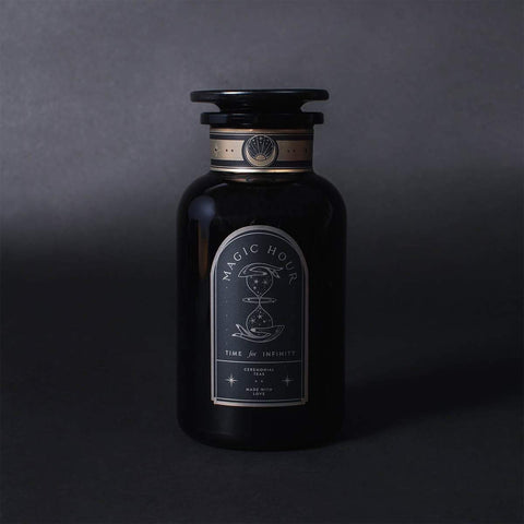 A dark, elegant apothecary-style bottle with a black label reading "Magic Hour," featuring minimalist design elements such as an hourglass and cosmic symbols. The bottle has a vintage, sophisticated appearance that evokes the purity of Club Magic Hour's Silver Moon White Tea, placed against a dark, gradient background.