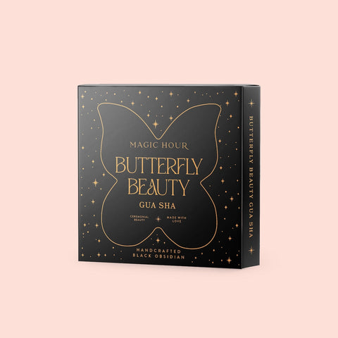 A black box with gold detailing and text on a light pink background. The box reads "Butterfly Beauty Gua Sha" with "Handcrafted Black Obsidian" at the bottom. The box, inspired by the elegance of Magic Hour, features a butterfly design and is decorated with small stars.