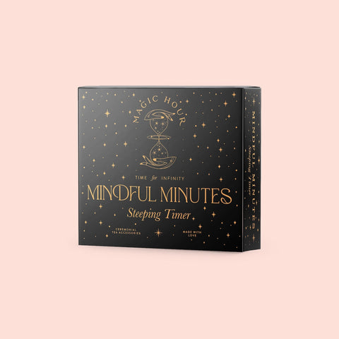 A rectangular, black box with gold stars and an hourglass design on the front. The text reads "MAGIC HOUR, Time for Infinity, Alibaba Mindful Minutes Tea Timer." The box claims it is "Ceremonial Tea Accessories" and is "Made with Love." The background is light pink.