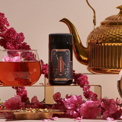 A tea setup with a jar labeled "The Magician by Magic Hour" surrounded by pink flowers, a gold teapot, and a glass cup of pink tea. Dried flower petals are scattered around, adding an elegant touch to the scene, hinting at the wellness benefits and transformative abilities of this enchanting brew.