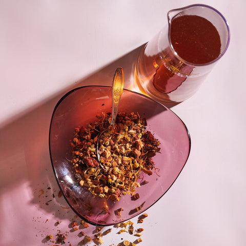 A heart-shaped, translucent pink bowl filled with loose leaf tea and a silver spoon rests on a surface. Next to the bowl is a clear glass pitcher, partially filled with red liquid, casting a shadow on the surface. The scene is lit with warm, diffuse light, invoking the transformative abilities of Magic Hour's The Magician wellness benefits.