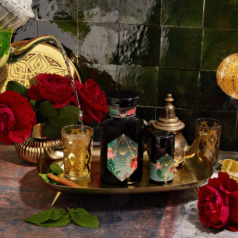 A decorative scene featuring a dark bottle with intricate golden and colorful designs, accompanied by a matching smaller container on an ornate golden tray showcases the opulence of Moonlight in Marrakesh: Spiced Mint Green Tea Case Packs by Magic Hour. Two lavishly adorned glasses are being filled with this exquisite Moroccan mint tea, while red roses and a teapot accentuate the setting.