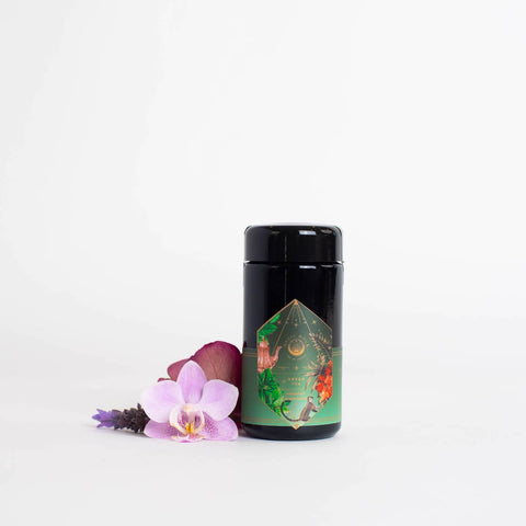 A Moonlight in Marrakesh: Spiced Mint Green Tea case pack by Magic Hour, with its black cylindrical container and decorative green and gold design, stands on a white surface. Surrounding it are pink and purple orchids, lavender flowers, and hints of Moroccan mint tea, adding a touch of nature and color to the scene.