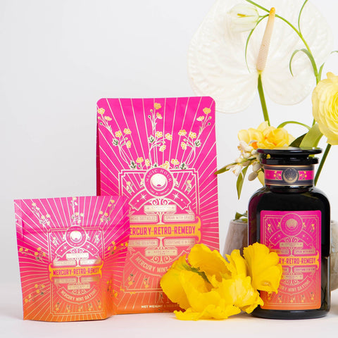 A vibrant display showcasing three Mercury Mint Daytime products in colorful pink and orange packaging adorned with astronomical designs. The set includes two pouches and a bottle of Vanilla Mint Lavender Jasmine Black Tea. They are surrounded by white and yellow flowers against a light background.