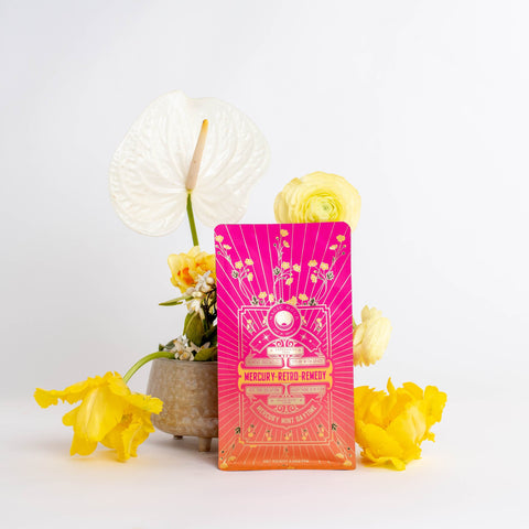 A vibrant pink and orange package labeled "Mercury Mint Daytime : Vanilla Mint Lavender Jasmine Black Tea" stands in front of a small vase containing white and yellow flowers. The backdrop is plain white, making the colors on the package and flowers pop, while hinting at the soothing presence of Organic Biodynamic Black Tea.