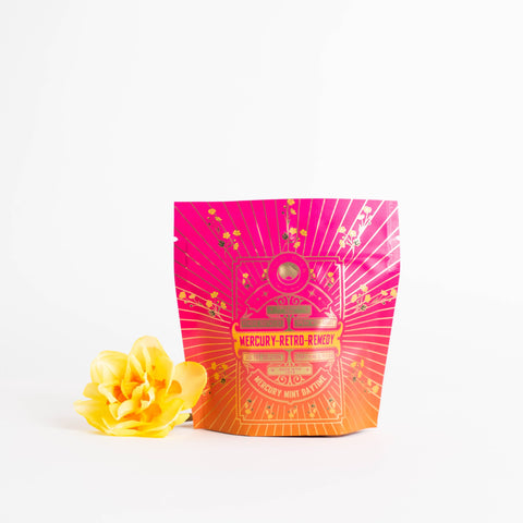 A vibrantly colored pouch labeled "Mercury Mint Daytime : Vanilla Mint Lavender Jasmine Black Tea" with decorative golden designs stands next to a yellow flower against a plain white background. The pouch, containing Organic Biodynamic Black Tea, transitions from pink at the top to orange at the bottom.