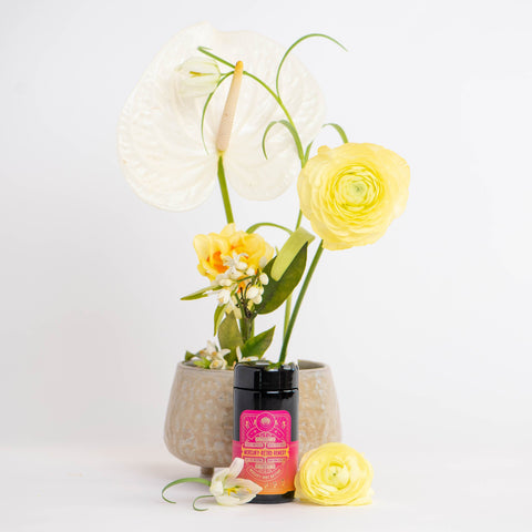 A small gray pot with yellow and white flowers on a white background sits behind a black container of Mercury Mint Daytime: Vanilla Mint Lavender Jasmine Black Tea, adorned with a pink label featuring graphic designs and text. A yellow flower lies next to the container on the surface.