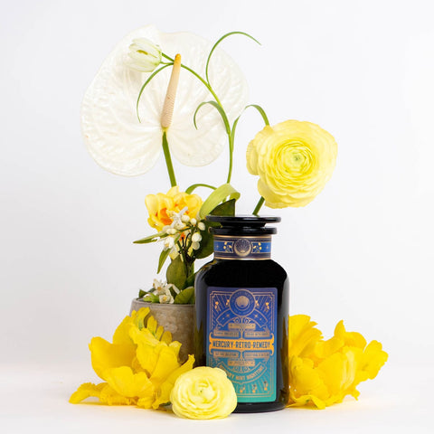 A black bottle with a blue and gold label reads "Mercury Mint Nighttime: Vanilla Mint Lavender Jasmine Tea- Caffeine Free!" The caffeine-free herbal blend is surrounded by an arrangement of yellow flowers and a white anthurium in a small pot. The backdrop is white, highlighting the colors of the flowers and the decorative bottle.