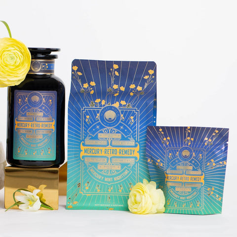 The image showcases a collection of three products labeled "Mercury Mint Nighttime: Vanilla Mint Lavender Jasmine Tea- Caffeine Free!" One item is a dark glass bottle with a gold-trimmed cap, another is a standing pouch, and the third is a small box, all adorned with a starry blue and green design. Arranged beside them are yellow and white flowers, highlighting the caffeine-free herbal blend featuring peppermint, vanilla mint, lavender, and jasmine.