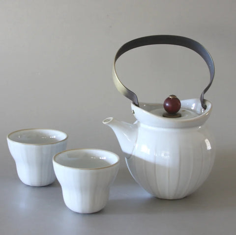 A white ceramic teapot with a black metal handle and a red knob on the lid is positioned next to two matching white teacups on a light-colored surface. This elegant Metal Handle Tea Set by Eastern Elm features a modern, minimalist design.