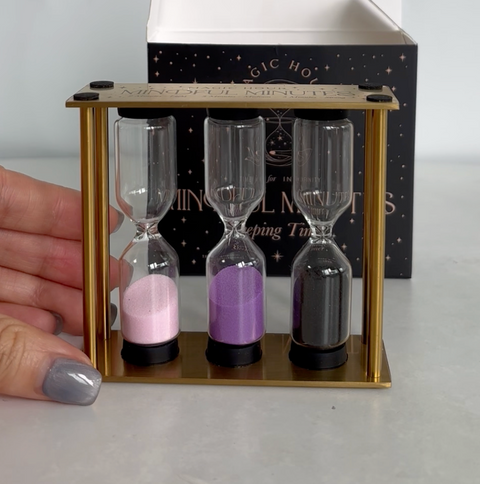 A person with gray-painted fingernails holds a golden frame with three hourglasses containing pink, purple, and black sand. Behind the frame, there is a partially open black box with small white stars and text that reads "Mindful Minutes Tea Timer by Alibaba.