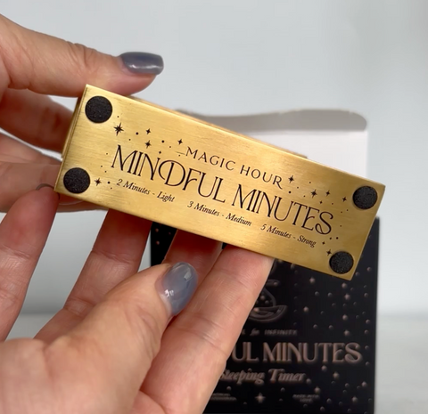 A person with gray nail polish holds a rectangular brass item labeled "Mindful Minutes Tea Timer." The item is marked with intervals: 2 Minutes - Light, 3 Minutes - Medium, 5 Minutes - Strong. The background includes a partially open box with similar branding from Alibaba.