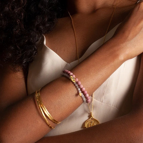 A person with curly hair wears a white spaghetti strap top, displaying gold bangles, the Rose Quartz Mini Hamsa Bracelet, and two beaded stretch bracelets in pink and purple. The arms are crossed, highlighting the jewelry against the background of light-colored clothing.