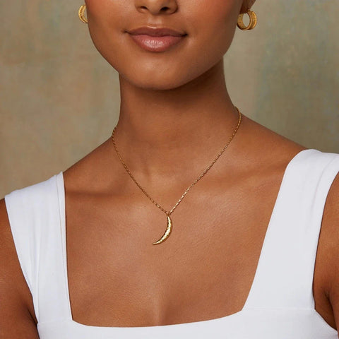 A person wearing a white sleeveless top, gold hoop earrings, and the Midnight Sky Pendant Moon Necklace featuring a celestial realm-inspired hammered 18kt crescent moon. The background is a plain, light, and slightly textured wall.