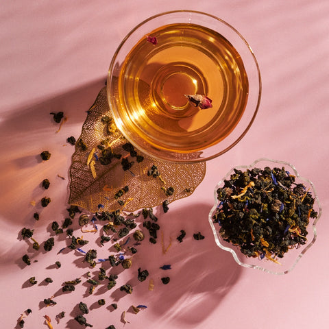 A transparent glass teacup filled with amber-colored Nuwa Tea for Intuition: Dragon Yuzu Oolong from Magic Hour sits on a pink surface. Next to it, a small ornate dish holds loose tea leaves. Some tea leaves are scattered around the dish, and the shadows create a textured pattern on the table.