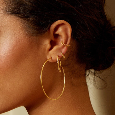 Close-up of an ear adorned with multiple gold earrings. The ear features an Ongoing Energy Large Hoop Earring by Magic Hour in the lower lobe, with two smaller hoops and a delicate ear cuff positioned on the upper ear cartilage. The hammered 18KT gold plate catches the light beautifully against the neutral background, highlighting the jewelry.