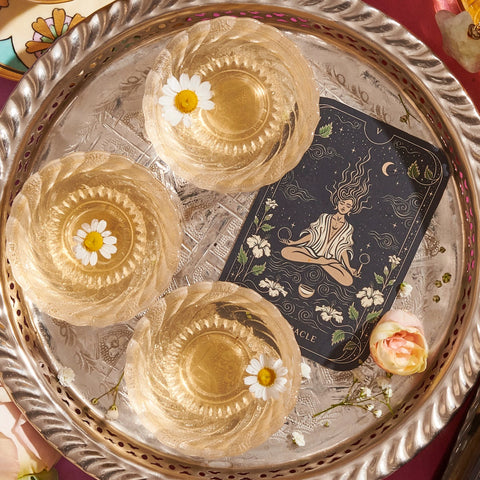 A decorative silver tray holds three translucent jelly desserts, each topped with a white daisy. Beside the desserts, there is a tarot card featuring a meditative figure surrounded by nature. A Magic Hour's "The Oracle: Lemon Lavender Tea for Supporting the Gut-Brain Axis" caffeine-free tea bag rests near the edge of the tray alongside a pink and white flower.