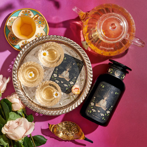 An elegant setup featuring a decorative tray with three glasses of champagne, a black bottle with an intricate label, Magic Hour's The Oracle: Lemon Lavender Tea for Supporting the Gut-Brain Axis in a clear teapot, a floral teacup, and a golden spoon with loose tea leaves, all arranged on a pink surface alongside Adaptogenic roots and white roses.