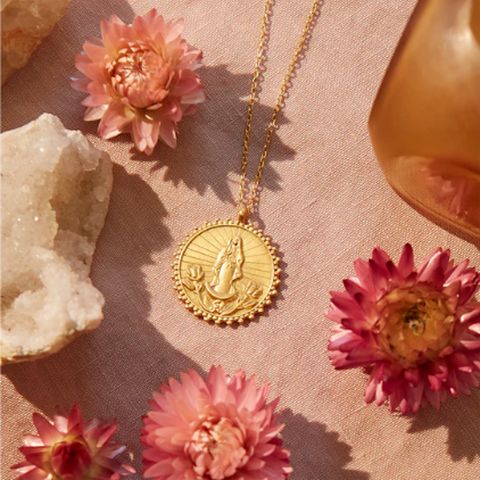 The Our Lady of Guadalupe, Divine Mother Necklace, featuring a gold chain and a circular pendant with an engraving of Our Lady of Guadalupe, is displayed on pink fabric. The necklace is elegantly surrounded by pink flowers, a rose-colored bottle, and a textured white stone, creating a soft and sophisticated aesthetic.